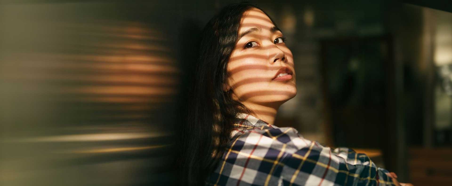 Woman with sunlight and shadow stripes on her face from Venetian blinds, gazing to the side.