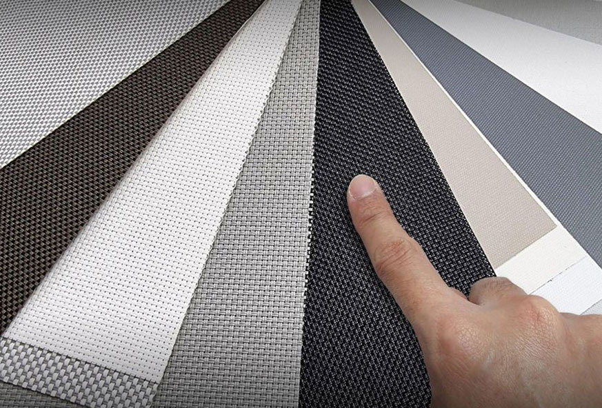 A finger points at a selection of vertical blind materials in various shades and textures.