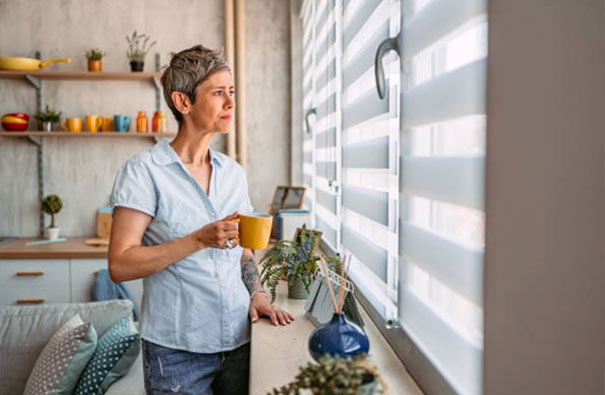 A woman gazes outside through her vision blinds with a cup in hand.