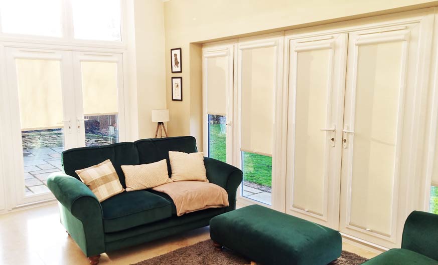 Cream Perfect Fit roller blinds on patio doors in a cosy lounge with a green sofa.